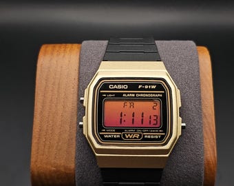 Peut inclure: Une montre numérique Casio F-91W dorée avec un bracelet noir. L'écran affiche l'heure 1:11:13 et le texte "ALARM CHRONOGRAPH". La montre est présentée sur un support en bois.