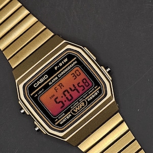 May include: Gold-toned Casio F-91W digital watch with a rectangular face and matching band. The watch face displays the time in red digits. Features include alarm, chronograph, and water resistance.