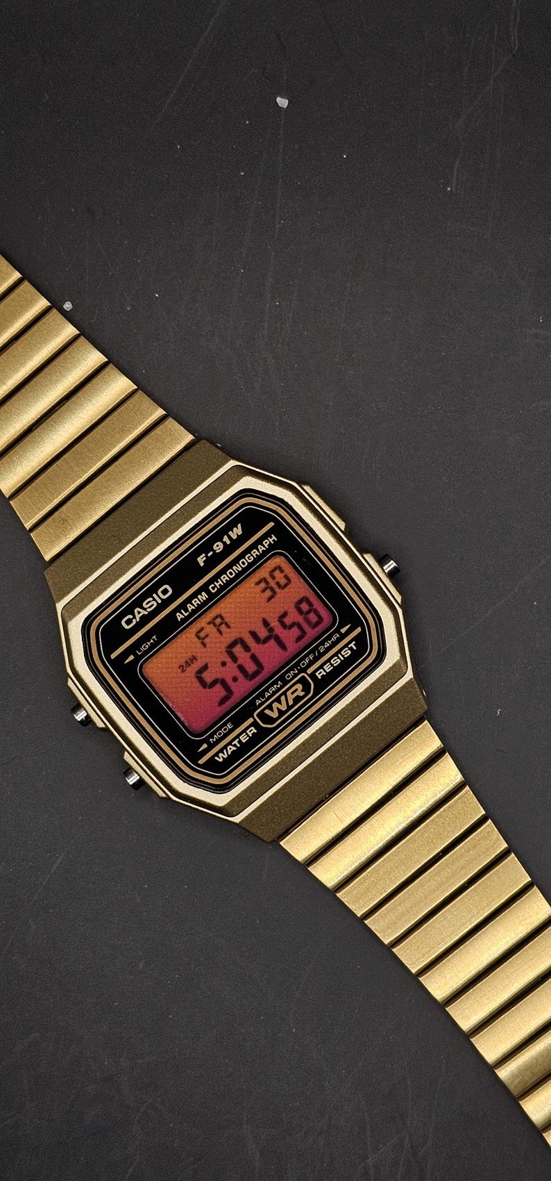 May include: Gold-toned Casio F-91W digital watch with a rectangular face and matching band. The watch face displays the time in red digits. Features include alarm, chronograph, and water resistance.