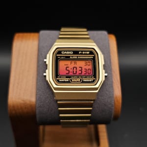 May include: Gold-toned Casio F-91W digital watch displaying the time 5:03:30 on a red digital display. The watch is on a wooden display stand with a gray cushion. The watch has the text "CASIO", "F-91W", "WATER RESIST" and "ALARM CHRONOGRAPH".