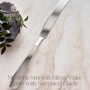 Può includere: Un moderno coltello da torta argentato con un manico liscio e una lama seghettata. Il coltello è esposto su una superficie di marmo. Il testo "Modern Smooth Silver Cake Knife with Serrated Blade" è visibile.