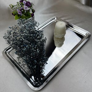 May include: A rectangular, silver-colored tray with a reflective surface. The tray holds a white candle and a bouquet of blue and white dried flowers. A small arrangement of purple flowers in a silver container is also present.