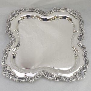 Pode incluir: Uma bandeja de servir prateada retangular com bordas ornamentadas e onduladas. A bandeja tem uma superfície reflexiva e uma borda decorativa com vieiras. O design apresenta intrincados padrões florais e foliares.