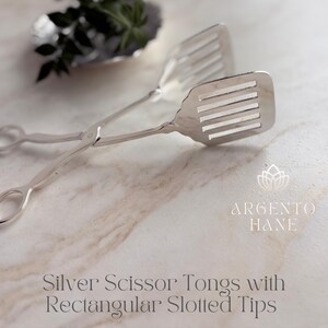 May include: Silver scissor tongs with rectangular slotted tips. The tongs are made of shiny silver metal. The tips have a slotted design. The text "Argento Hane" is in the image.