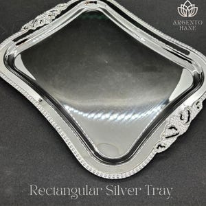 May include: A rectangular silver tray with ornate detailing around the edges. The tray has a reflective surface and the text "Rectangular Silver Tray" is displayed at the bottom. The tray is from Argento Hane.
