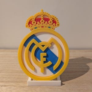 Peut inclure: Un emblème imprimé en 3D avec un design circulaire jaune et bleu avec les lettres "RMC" et une couronne. L'emblème est monté sur une base blanche. La couronne est jaune et rouge.