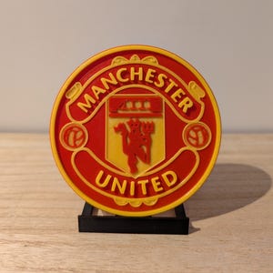 Puede incluir: Logotipo del Manchester United en rojo y amarillo, con las palabras "Manchester United" en un diseño circular. El logotipo incluye un diablo rojo y está montado sobre un soporte negro.