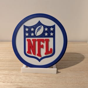 Peut inclure: Logo NFL imprimé en 3D, de forme circulaire. Le logo présente un fond blanc avec un bord bleu, les lettres NFL en rouge, un ballon de football et des étoiles en blanc et bleu. Il est présenté sur un support blanc.