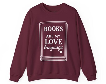 May include: Burgundy-colored sweatshirt with the text "BOOKS ARE MY LOVE language" in white, inside a book outline. The sweatshirt has long sleeves and a crew neck. A small white heart is below the word "language".