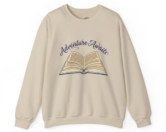 May include: A cream-colored sweatshirt with a crew neck. The front features a blue illustration of an open book with the words "Adventure Awaits" in a script font above the book. The sweatshirt is made of a soft material.