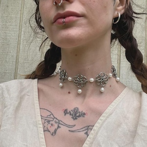 Puede incluir: Collar gargantilla plateado con diseños florales y perlas falsas colgantes. El collar se lleva alrededor del cuello. Una joya delicada.