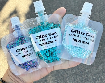 May include: Three pouches of Glittr Goo hair glitter gel with white screw caps. The labels read Mermaid, Laguna Glow, and Pastel Blue. Each pouch contains 1.55 oz (55 g) of glitter gel in purple, teal, and light blue colors with various shapes.