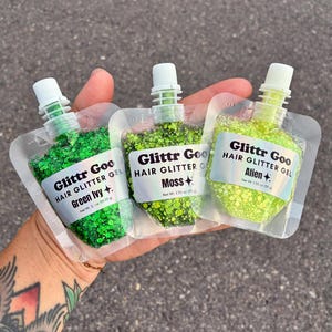 May include: Three clear plastic pouches of Glittr Goo hair glitter gel in shades Green Ivy, Moss, and Alien. Each pouch features a white label with black text and a small star icon, with a net weight of 1.35 oz (38 g) printed on each label.