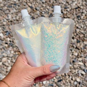 May include: Two clear plastic pouches with white screw-top caps, filled with iridescent gel. One pouch contains fine, shimmering white glitter, while the other contains larger, chunky, multi-colored iridescent glitter flakes.
