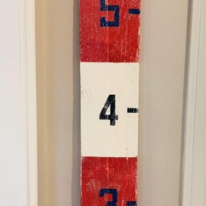 May include: A wooden nautical marker painted in red, white, and blue. The top section has the letters "I", "A", "E", and "5" in a diamond shape. Numbers 2 through 5 are displayed in blue, with dashes.