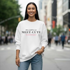 Puede incluir: Sudadera blanca con el texto negro "this could be our MEET-CUTE" y "Your storybook romance" en rosa. La sudadera se combina con unos vaqueros azules. La imagen está tomada al aire libre.