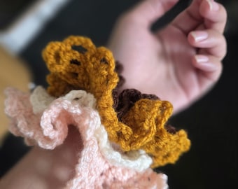 May include: A layered crochet hair accessory featuring three distinct ruffled bands in shades of soft pink, cream, and mustard yellow. The yarn is worked in an open, scalloped stitch pattern that creates a voluminous, textured appearance.