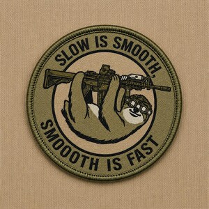 May include: A circular olive green embroidered patch featuring a sloth wearing goggles and holding a rifle. The text SLOW IS SMOOTH, SMOOTH IS FAST is printed in black capital letters along the border of the patch against a tan background.