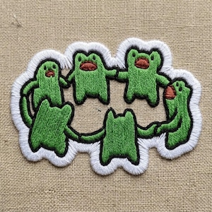 May include: Embroidered patch featuring five green cartoon frogs holding hands in a circle. Each frog has a red mouth and is outlined in black. The patch has a white border.