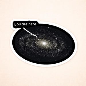 May include: A sticker featuring a stylized galaxy illustration with a bright center and numerous stars. The text "you are here" is in a speech bubble pointing to the galaxy. The sticker has a white border and is set against a light beige background.