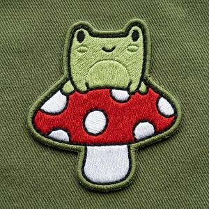May include: Embroidered patch featuring a green frog sitting atop a red mushroom with white spots. The patch has a black border and is set against a green fabric background. This patch is ideal for adding to clothing or accessories.