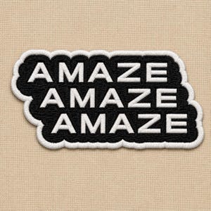 May include: A black embroidered patch with a white border featuring the word AMAZE repeated three times in a stacked, staggered layout. The white text is in a bold, sans-serif font, and the patch is set against a light beige fabric background.