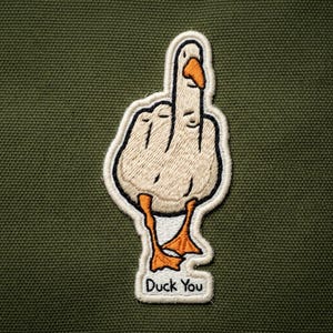 May include: Embroidered patch featuring a goose giving the middle finger. The goose is white with orange beak and feet, outlined in black. The text "Duck You" is at the bottom in black. The patch is on a green background.