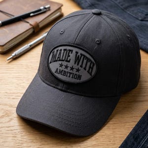 May include: A charcoal gray baseball cap with a curved brim. The cap features a patch with the words "MADE WITH AMBITION" and four stars. The hat is made of a durable fabric and has a button on top.