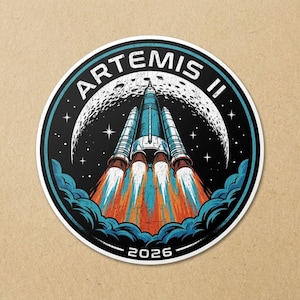 May include: Circular sticker featuring a rocket launching into space, with the text "ARTEMIS II" in white. The rocket is blue and white, with orange and red flames. The background includes a moon, stars, and the year "2026".