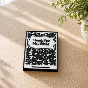 May include: A black and white notebook-style item with the text "Thank You Mr. While" on the cover. The design resembles a classic composition book. The item is on a light-coloured wooden surface.