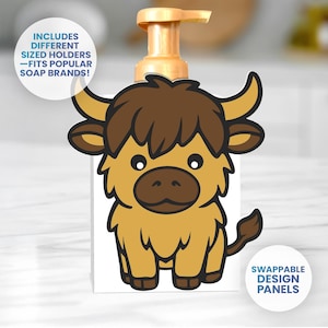 Könnte beinhalten: Ein weißer Seifenspender mit einem Cartoon-Yak-Design. Das Yak ist braun und hellbraun mit schwarzen Umrissen. Der Spender hat eine goldene Pumpe und den Text "INCLUDES DIFFERENT SIZED HOLDERS - FITS POPULAR SOAP BRANDS!" und "SWAPPABLE DESIGN PANELS".