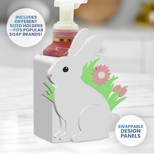 May include: A white soap dispenser holder with a bunny design, green grass, and pink flowers. The holder has a rectangular base and includes the text "INCLUDES DIFFERENT SIZED HOLDERS - FITS POPULAR SOAP BRANDS!" and "SWAPPABLE DESIGN PANELS".