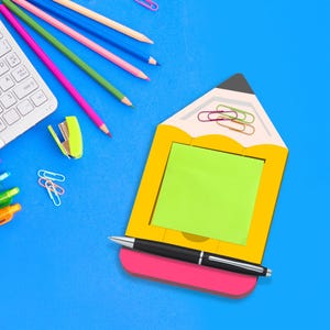 May include: A yellow pencil-shaped memo pad with a green sticky note, a black pen, and a pink eraser. Colored pencils, paper clips, and a keyboard are scattered on a blue surface. A lime green pencil sharpener is also visible.
