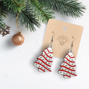 May include: A pair of Christmas tree-shaped earrings. The earrings are white with red and green accents, resembling a frosted cookie. The earrings hang from silver-colored hooks. The background includes a sprig of evergreen and a gold ornament.