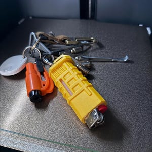 May include: A set of keys on a key ring, featuring a yellow lighter, an orange tool, and a white circular tag. The keys are diverse in shape and size, with a leather key fob attached.