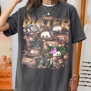 Puede incluir: Camiseta gris oscuro con un dise&ntilde;o del personaje Tow Mater de la pel&iacute;cula Cars. El dise&ntilde;o incluye varios personajes de coches animados y el texto "Tow Mater" en letras doradas. La camiseta tiene un aspecto vintage y desgastado.