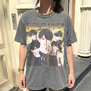 Può includere: Una maglietta grigia a maniche corte con una grafica di personaggi in stile anime. Il testo "GIRL DINNER" è stampato in giallo e arancione. La maglietta è indossata da una persona.