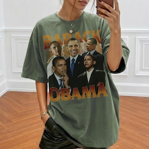 May include: Olive green t-shirt featuring a graphic of Barack Obama in various poses, with the name "OBAMA" in a gradient orange and gold font. The word "BARACK" is above the images. The shirt has a vintage, slightly faded look.