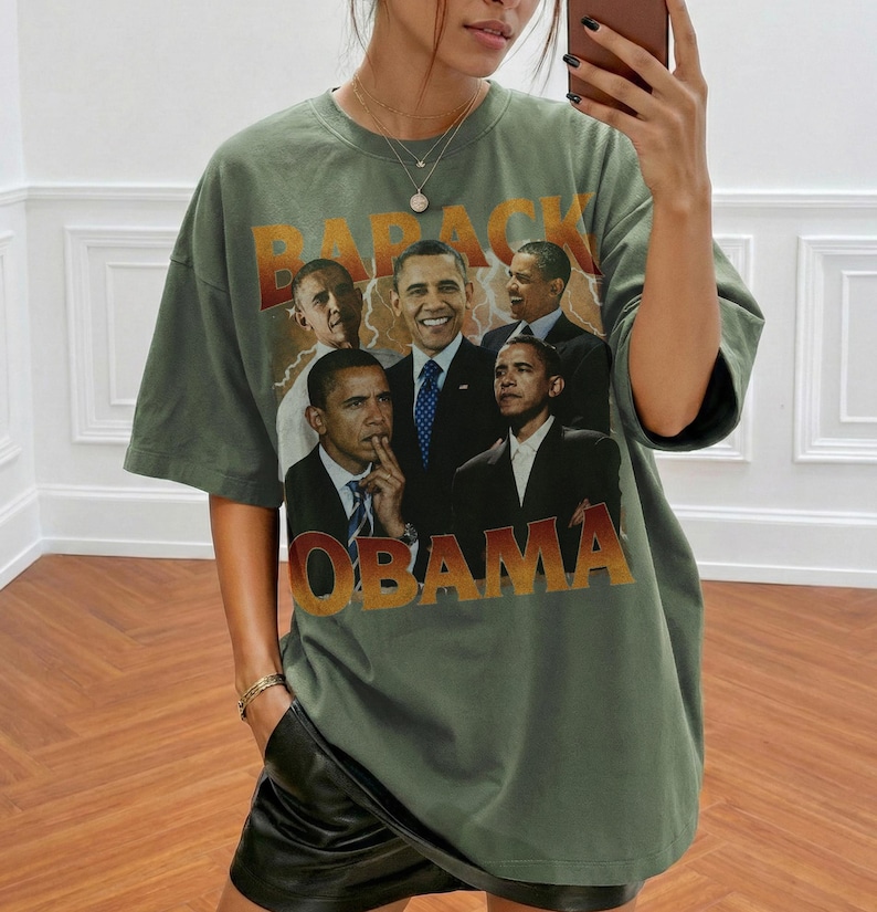 May include: Olive green t-shirt featuring a graphic of Barack Obama in various poses, with the name "OBAMA" in a gradient orange and gold font. The word "BARACK" is above the images. The shirt has a vintage, slightly faded look.