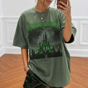 May include: Olive green t-shirt with a graphic of a castle and the word "Disneyland" in a green, gothic font. The shirt has short sleeves and a relaxed fit. The graphic features fireworks and a dark, ominous sky.