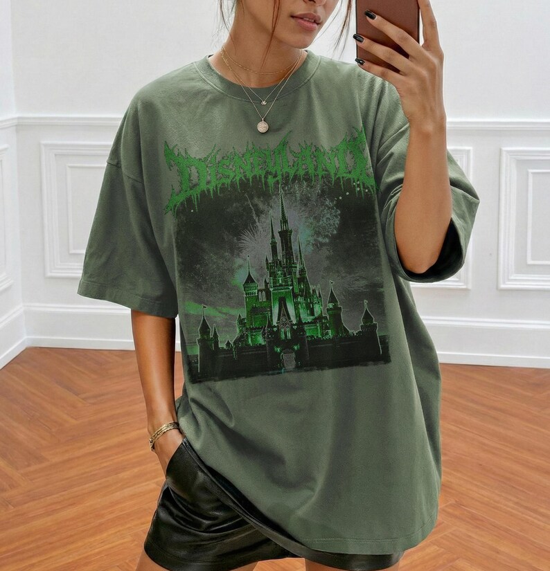 May include: Olive green t-shirt with a graphic of a castle and the word "Disneyland" in a green, gothic font. The shirt has short sleeves and a relaxed fit. The graphic features fireworks and a dark, ominous sky.