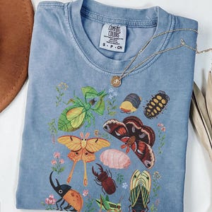 May include: A blue short-sleeved t-shirt featuring a graphic print of various insects and small flowers. The shirt has a Comfort Colors brand label at the neck, which reads S-P-CH, 100% cotton, and made in Honduras. A gold-toned necklace rests on the fabric.