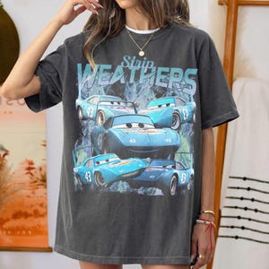 May include: Dark gray t-shirt featuring a graphic of blue race cars with the number 43. The text "Strip Weathers" is above the cars. The shirt has a vintage, worn look.