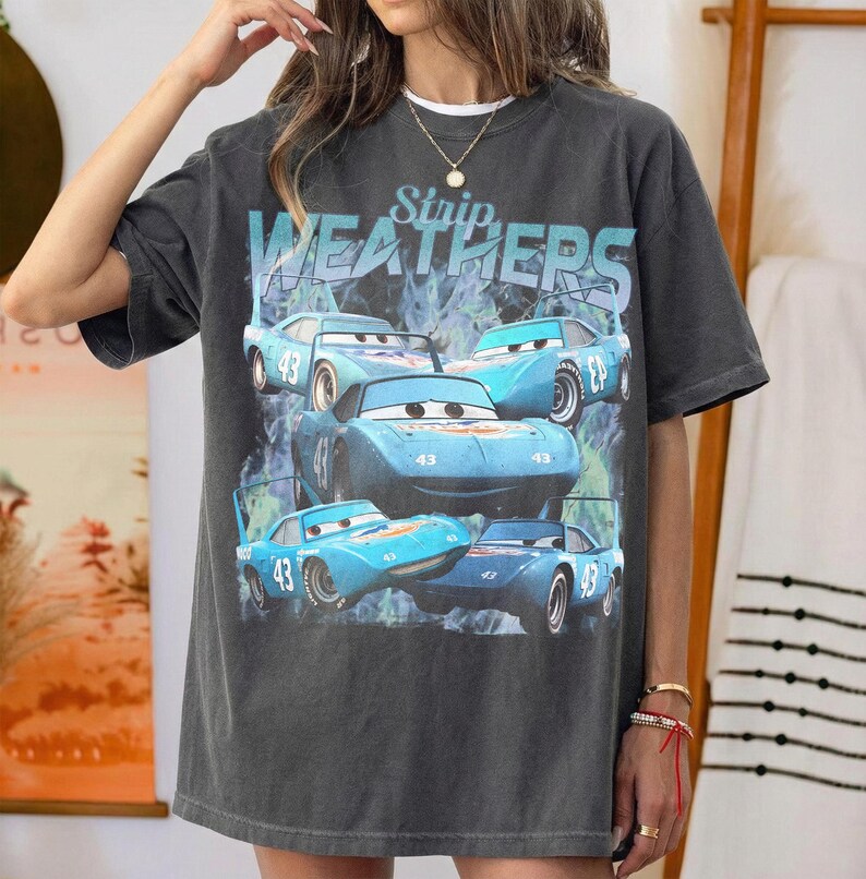 May include: Dark gray t-shirt featuring a graphic of blue race cars with the number 43. The text "Strip Weathers" is above the cars. The shirt has a vintage, worn look.