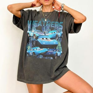 May include: Dark gray t-shirt featuring a graphic of blue race cars with the number 43 and the text "Strip Weathers". The cars are set against a blue and green background. The shirt has short sleeves and a relaxed fit.