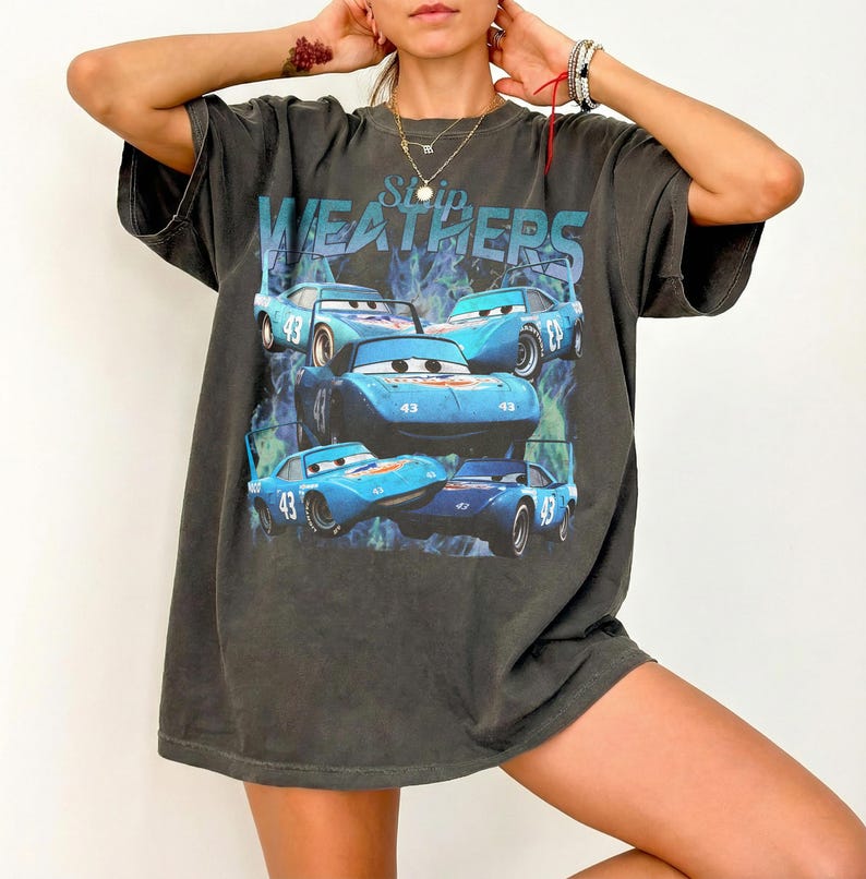 May include: Dark gray t-shirt featuring a graphic of blue race cars with the number 43 and the text "Strip Weathers". The cars are set against a blue and green background. The shirt has short sleeves and a relaxed fit.