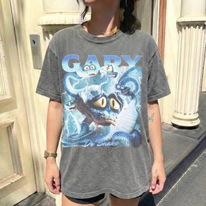 Pode incluir: Camiseta cinza com uma ilustra&ccedil;&atilde;o de desenho animado de uma criatura semelhante a uma cobra e o nome "GARY" em azul. O design inclui raios azuis e as palavras "De Snake".
