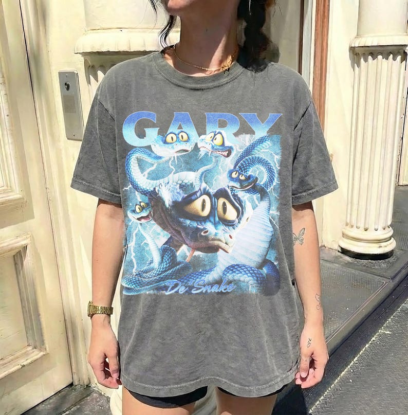 Pode incluir: Camiseta cinza com uma ilustra&ccedil;&atilde;o de desenho animado de uma criatura semelhante a uma cobra e o nome "GARY" em azul. O design inclui raios azuis e as palavras "De Snake".