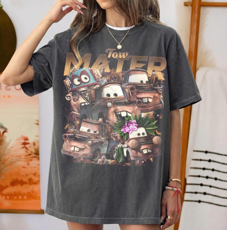 Puede incluir: Camiseta gris oscuro con un dise&ntilde;o del personaje Tow Mater de la pel&iacute;cula Cars. El dise&ntilde;o incluye varios personajes de coches animados y el texto "Tow Mater" en letras doradas. La camiseta tiene un aspecto vintage y desgastado.