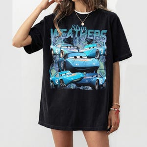 May include: Black t-shirt featuring a graphic of blue race cars with the number 43. The text "Strip Weathers" is above the cars. The shirt has a relaxed fit and short sleeves.